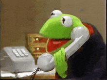 kermit the frog is talking on a telephone while wearing a green shirt .