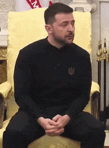 a man is sitting in a chair with his hands folded in front of a flag that says ny
