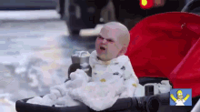 a baby is sitting in a stroller with a homer simpson logo on the bottom right corner .