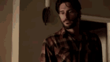 a man with a beard wearing a plaid shirt is standing in a room .