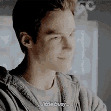 a young man is smiling and saying `` little busy '' while wearing a grey hoodie .