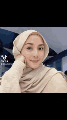 a woman wearing a hijab and a white sweater has a tiktok sticker on her face
