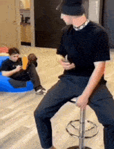 a man is sitting on a bar stool while another man sits on a bean bag chair and looks at his phone .