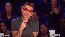 a man wearing red glasses is sitting in front of a microphone with a nbc logo on the cup in front of him