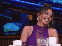 a woman in a purple dress is laughing while sitting at a table with coffee cups .