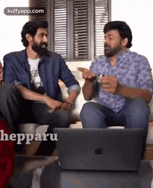 two men are sitting on a couch talking to each other and looking at a laptop .