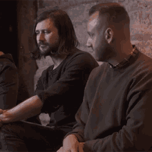 a man with long hair and a beard sits next to another man in a brown sweater