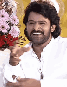 a man with a beard and mustache is smiling and pointing at the camera in front of a bouquet of pink flowers .