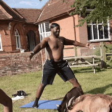 a shirtless man stands on a yoga mat surrounded by goats in front of a brick building