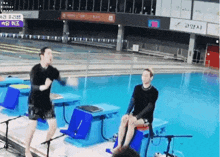 a man is standing in front of a swimming pool while another man sits on a bench next to him .