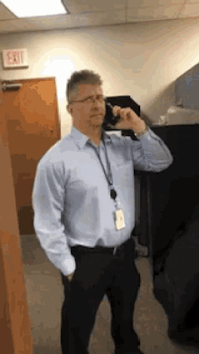 a man in a light blue shirt and black pants is talking on a cell phone in an office .