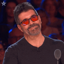 a man wearing red glasses and a black sweater smiles in front of a crowd