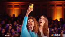 two women taking a selfie in front of a crowd with the nbc logo on the bottom right