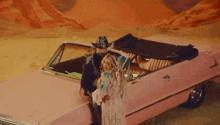 a man and a woman are standing next to a pink car . the woman is wearing a cowboy hat .