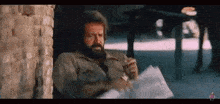 a man with a beard is sitting in a chair smoking a cigarette and reading a book .