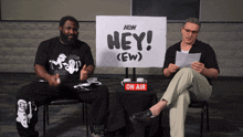 two men are sitting in front of a sign that says hey ( ew )