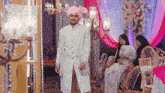 a man in a white suit and a pink turban is standing in a room with a woman sitting on a couch .