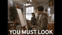 a man in a suit is being hugged by a woman in front of an easel with the words " you must look " above him