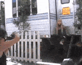 a woman is taking a picture of a woman standing next to a trailer behind a white picket fence .