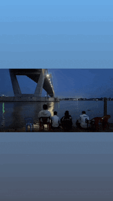 a group of people sitting on the shore of a body of water looking at a bridge