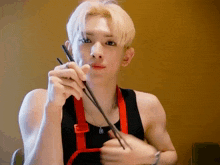 a young man wearing a black tank top and an apron is eating with chopsticks .