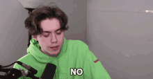 a young man in a green hoodie is sitting in front of a microphone and says no .