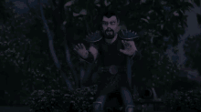 a man with a beard and spikes on his arms is sitting in the woods at night .