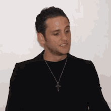 a man wearing a black jacket and a cross necklace is standing in front of a white wall .