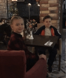 a boy and a girl sit at a table with a vase of flowers on it