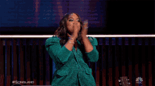 a woman in a green jacket applauds in front of a guitar and a nbc logo
