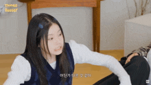 a girl in a blue vest is sitting on the floor talking to another girl in korean
