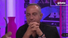 a man is sitting at a table with his hands folded in front of a purple background .