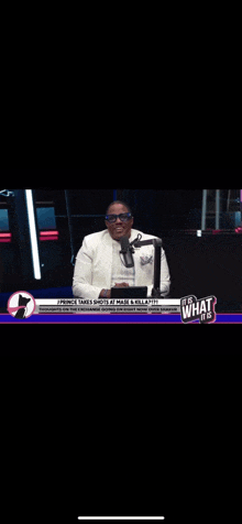 a man in a white suit stands in front of a microphone with the words it 's what it 's on the bottom right