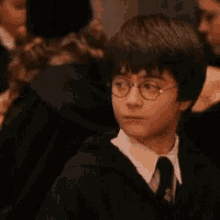 harry potter is wearing glasses and a tie and is sitting in a crowd of people .