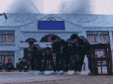a group of people are squatting in front of a building with a blue sign on the top that says ' squid game '