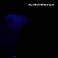 a man in a blue shirt is holding a helmet in front of a mumbaiindians.com banner