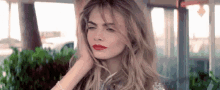 a woman with long blonde hair and red lipstick is sitting in front of a window .
