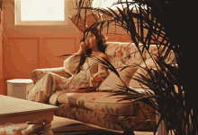 a woman sits on a floral couch in a living room with a plant in the foreground