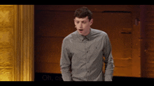 a man in a grey polo shirt stands in front of a microphone with the words oh can you believe it behind him