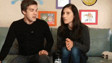 a man and a woman are sitting on a couch with a picture on the wall that says " get me "