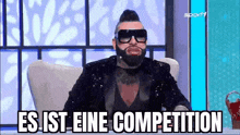 a man with a beard and sunglasses is sitting in a chair with the words `` es ist eine competition '' written on the screen .