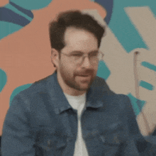 a man wearing glasses and a denim jacket is pointing to his mouth