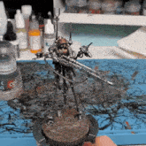 a person is holding a miniature of a robot with a sniper rifle in front of a bottle of colossus paint