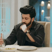 a man with a beard is sitting at a table drinking from a white cup .