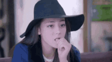 a woman wearing a hat and a blue jacket is sitting at a table with her hand on her chin .