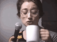 a woman wearing glasses is drinking from a white cup while holding a microphone .