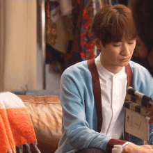 a man in a blue cardigan sits on a couch looking at a clapper board that says " fly "