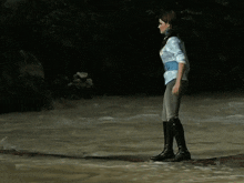 a woman in a blue shirt and black boots is walking across a stream