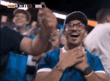 a man wearing glasses and a blue shirt applauds in front of a screen that says goal
