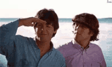 two men are standing next to each other on the beach and one of them is covering his eyes with his hand .